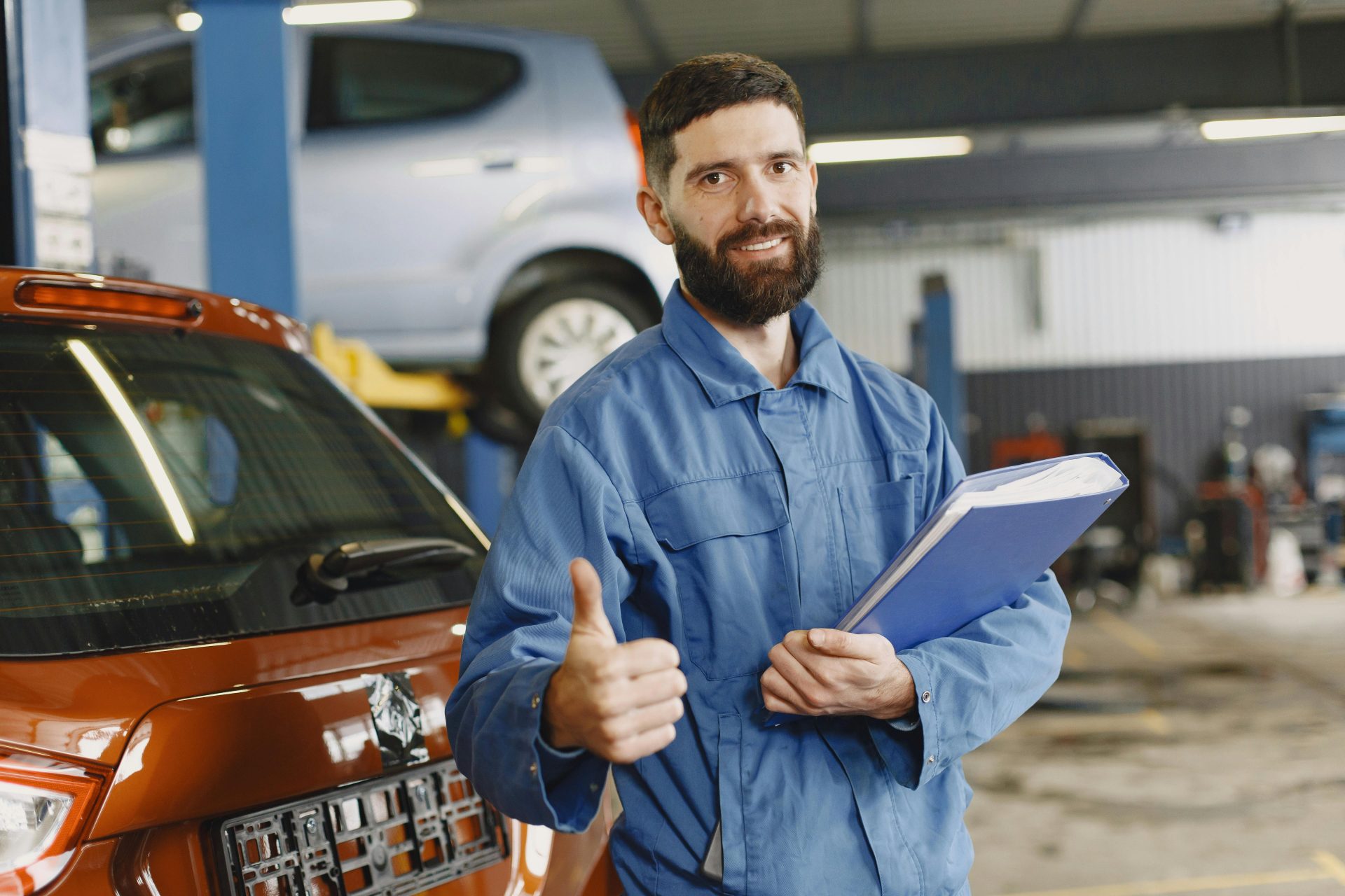 Automechaniker im blauen Overall mit Klemmbrett, der in der Werkstatt den Daumen nach oben zeigt.